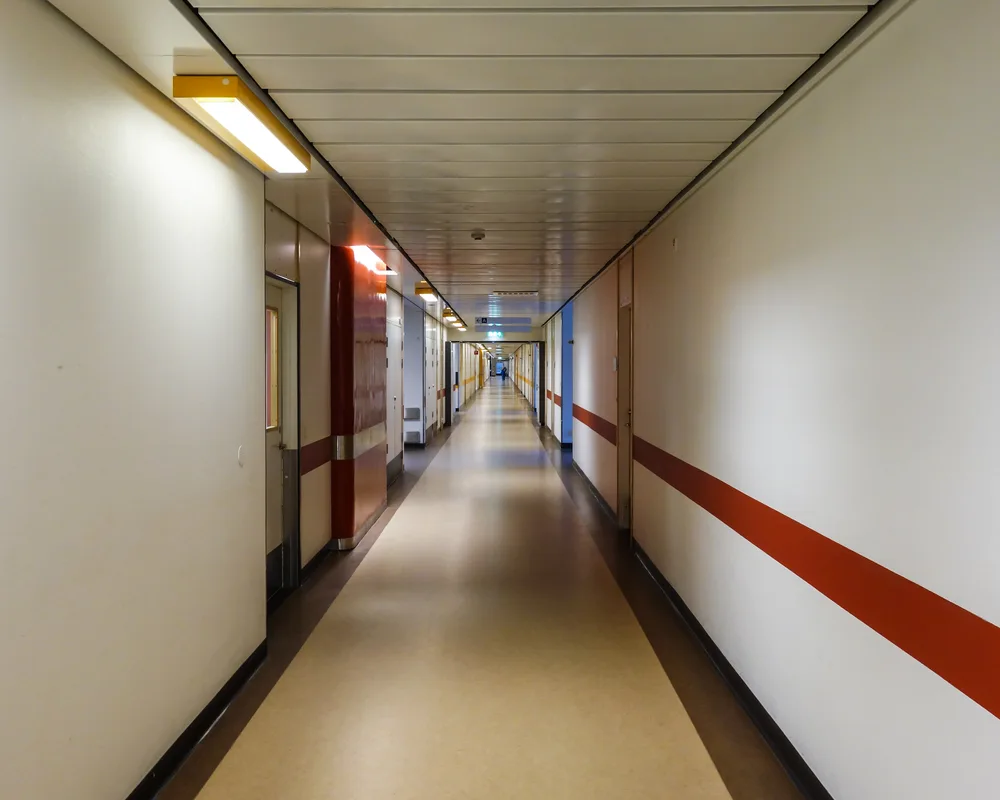 A long, empty, brightly lit hospital corridor with a red accent stripe along the wall.